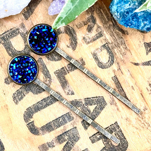 All Up In The Hair | Online Accessory Boutique Located in Mooresville, NC | Two dark blue druzy bobby pins on a wood background with black lettering. There are crystals and ivy leaves at the top of the image.