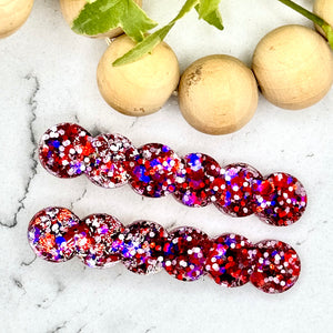 All Up In The Hair | Online Accessory Boutique Located in Mooresville, NC | Two red, blue, and silver barrettes laying on a white marble background. At the top of the picture is a wood bead garland and ivy leaves.
