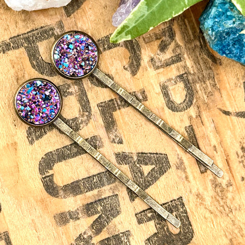 All Up In The Hair | Online Accessory Boutique Located in Mooresville, NC | Two Rainbow Druzy Bobby Pins laying on a wood background with black lettering. There are crystals and ivy leaves at the top of the image.