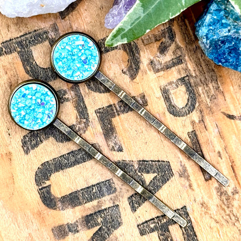 All Up In The Hair | Online Accessory Boutique Located in Mooresville, NC | Two light blue druzy bobby pins on a wood background with black lettering. There are crystals and ivy leaves at the top of the image.