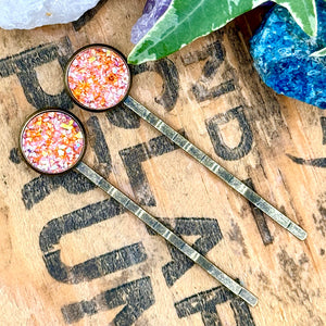 All Up In The Hair | Online Accessory Boutique Located in Mooresville, NC | Two orange druzy bobby pins laying diagonally on a wood background with black lettering. There are crystals and ivy leaves at the top of the image.