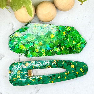 All Up In The Hair | Online Accessory Boutique Located in Mooresville, NC | An angular barrette filled with four leaf clover glitter and a teardrop shape barrette made with green and gold glitter laying on a white marble background. At the top of the image is a wood bead garland and ivy leaves.