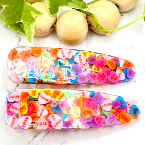All Up In The Hair | Online Accessory Boutique Located in Mooresville, NC | Two teardrop shaped barrettes filled with colorful hearts on a white marble background. At the top of the image is a wood bead garland and ivy leaves.