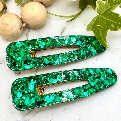 All Up In The Hair | Online Accessory Boutique Located in Mooresville, NC | Two hollow teardrop shaped barrette made with green glitter laying on a white marble background. At the top of the image is a wood bead garland and ivy leaves.