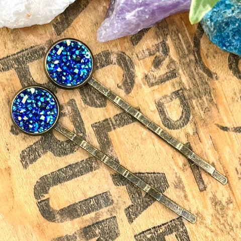 All Up In The Hair | Online Accessory Boutique Located in Mooresville, NC | Two blue druzy bobby pins laying diagonally on a wood background with black lettering. There are crystals and ivy leaves at the top of the image.