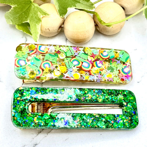 All Up In The Hair | Online Accessory Boutique Located in Mooresville, NC | Two rectangular barrettes on a white marble background. The top barrette is filled with "lucky charms" like rainbows, pots of gold, stars, leprechaun hats, etc. The bottom barrette is made with a bright green glitter. At the top of the image is a wood bead garland and ivy leaves.