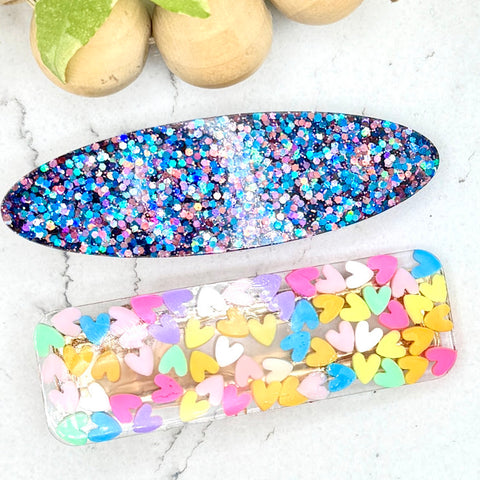 All Up In The Hair | Online Accessory Boutique Located in Mooresville, NC | An oval barrette made with blue and pink glitter and a rectangle barrette filled with pastel hearts laying on a white marble background. At the top of the image is a wood bead garland and ivy leaves.