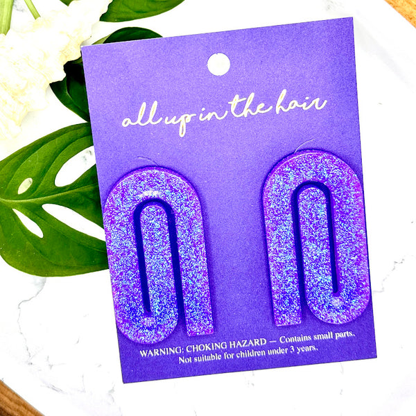 All Up In The Hair | Online Accessory Boutique Located in Mooresville, NC | Two purple paperclips on an indigo packaging card. The card is laying on monstera leaves on a white background.