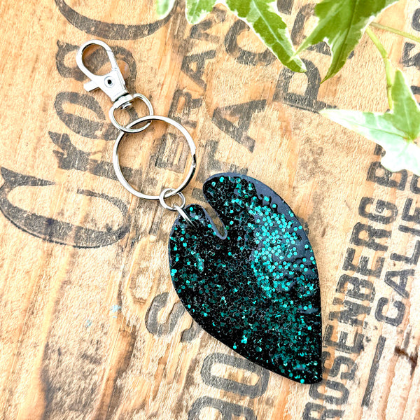 All Up In The Hair | Online Accessory Boutique Located in Mooresville, NC | Back of our Alocasia Leaf Keychain on a wood background with black lettering. There are ivy leaves next to the keychain.