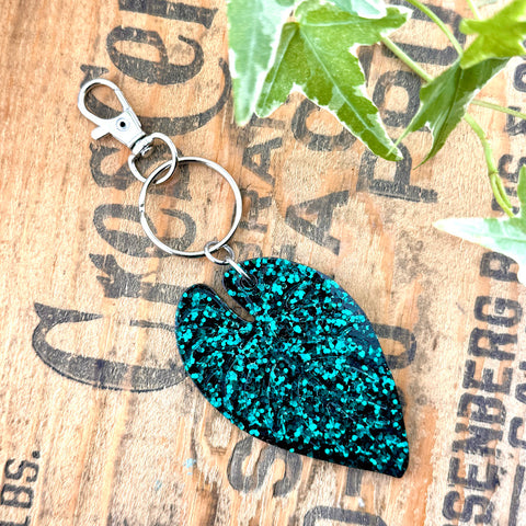 All Up In The Hair | Online Accessory Boutique Located in Mooresville, NC | A green glitter Alocasia Leaf Keychain on a wood background with black lettering. There are ivy leaves next to the keychain.