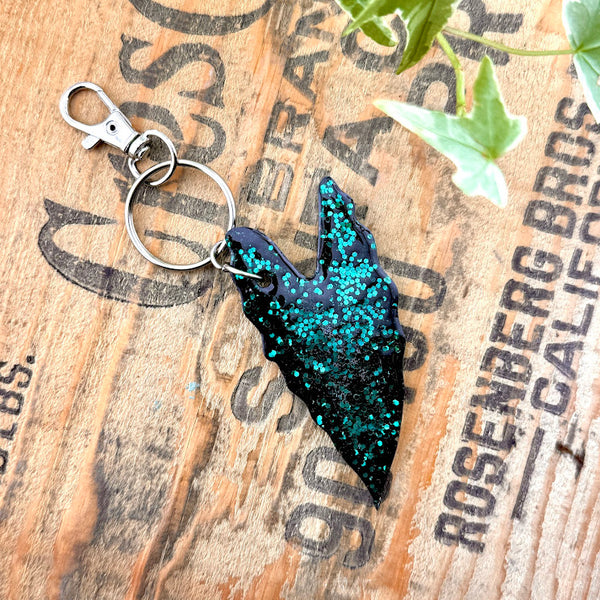 All Up In The Hair | Online Accessory Boutique Located in Mooresville, NC | Back of our Alocasia Leaf Keychain on a wood background with black lettering. There are ivy leaves next to the keychain.