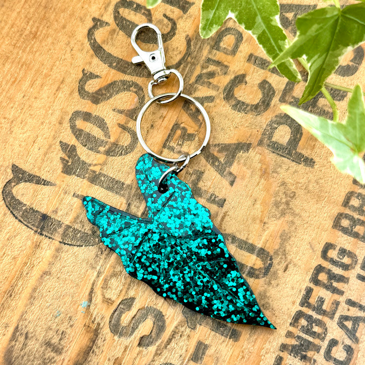 All Up In The Hair | Online Accessory Boutique Located in Mooresville, NC | A green glitter Alocasia Leaf Keychain on a wood background with black lettering. There are ivy leaves next to the keychain.