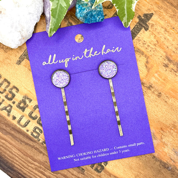 All Up In The Hair | Online Accessory Boutique Located in Mooresville, NC | An All Up In The Hair branded packaging card laying diagonally on a grey background surrounded by colorful glitter. On the card its two amethyst druzy bobby pins.