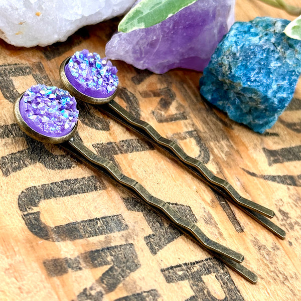 All Up In The Hair | Online Accessory Boutique Located in Mooresville, NC | Side view of our Amethyst Druzy Bobby Pins laying on a wood background with black lettering. There are crystals and ivy leaves at the top of the image.