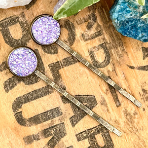 All Up In The Hair | Online Accessory Boutique Located in Mooresville, NC | Two purple druzy bobby pins laying on a wood background with black lettering. There are crystals and ivy leaves at the top of the image.