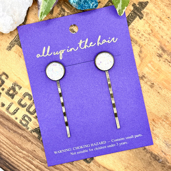 All Up In The Hair | Online Accessory Boutique Located in Mooresville, NC | Our Angel Aura Quartz Druzy Bobby Pins on an indigo colored, All Up In The Hair branded packaging card. The card is laying on a wood background with black lettering. There are crystals and ivy leaves at the top of the image.