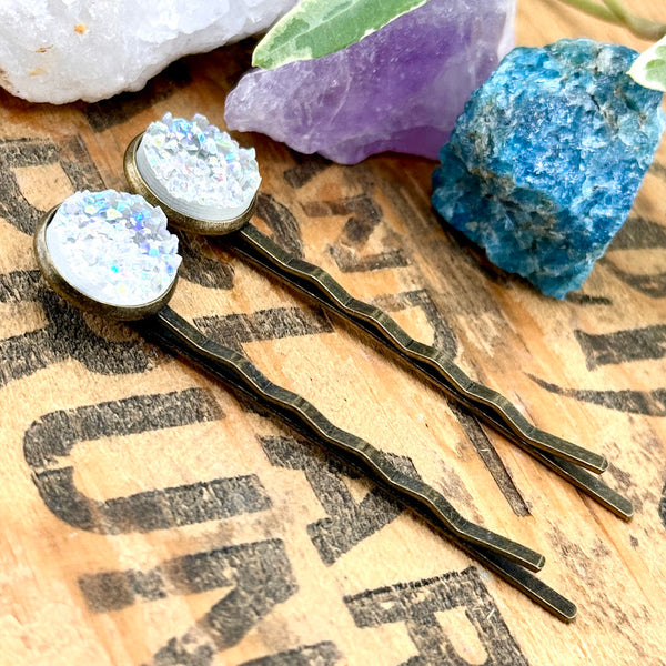 All Up In The Hair | Online Accessory Boutique Located in Mooresville, NC | Side view of our Angel Aura Quartz Druzy Bobby Pins laying on a wood background with black lettering. There are crystals and ivy leaves at the top of the image.