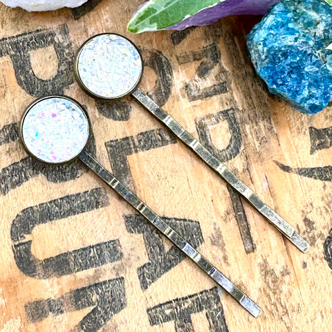 All Up In The Hair | Online Accessory Boutique Located in Mooresville, NC | Two iridescent druzy bobby pins laying diagonally on a grey background, surrounded by colorful glitter.