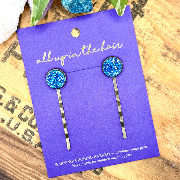 All Up In The Hair | Online Accessory Boutique Located in Mooresville, NC | An All Up In The Hair branded packaging card laying on a grey background, surrounded by colorful glitter. On the card is two apatite druzy bobby pins.