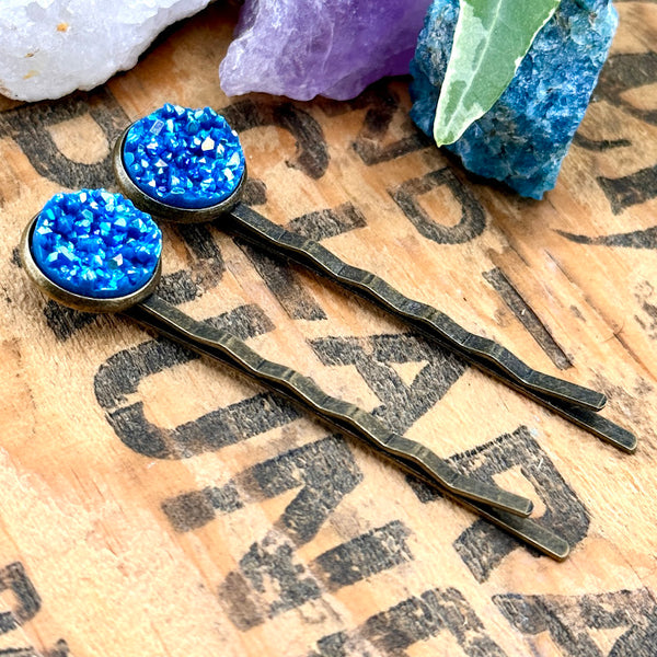 All Up In The Hair | Online Accessory Boutique Located in Mooresville, NC | Side view of our Apatite Druzy Bobby Pins laying on a wood background with black lettering. There are crystals and ivy leaves at the top of the image.