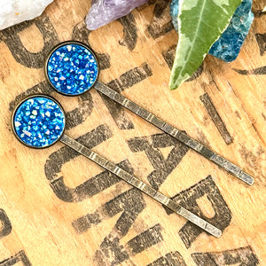 All Up In The Hair | Online Accessory Boutique Located in Mooresville, NC | Two blue druzy bobby pins laying on a wood background with black lettering. There are crystals and ivy leaves at the top of the image.