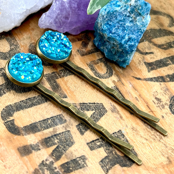 All Up In The Hair | Online Accessory Boutique Located in Mooresville, NC | Two Aquamarine Druzy Bobby Pins laying on a wood background with black lettering. There are crystals and ivy leaves at the top of the image.