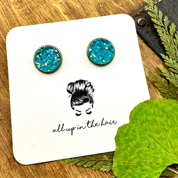 All Up In The Hair | Online Accessory Boutique Located in Mooresville, NC | Two Aquamarine Druzy set in a bronze stud setting. The earrings are on a white, All Up In The Hair branded packaging card. The card is laying on a wood background, alongside green leaves.