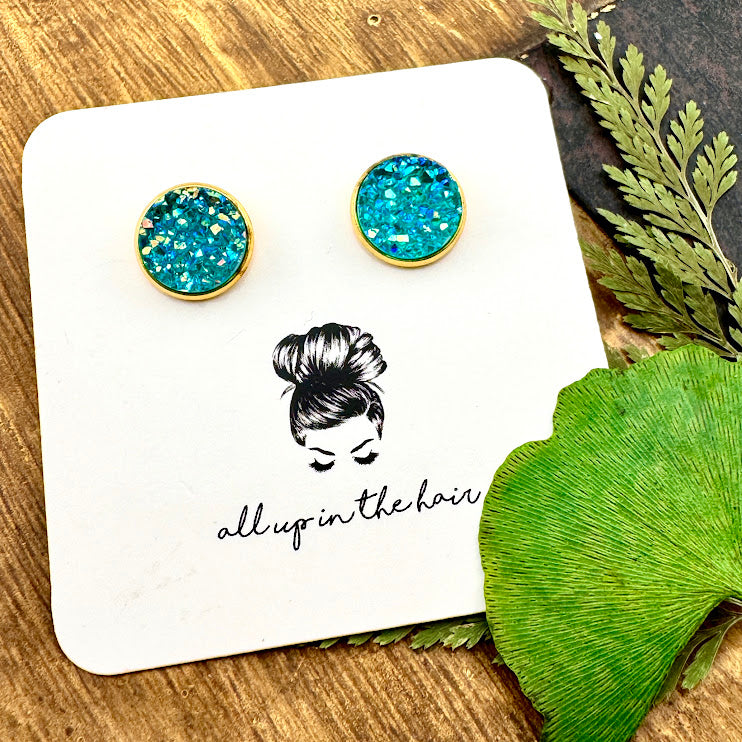 All Up In The Hair | Online Accessory Boutique Located in Mooresville, NC | Two Aquamarine Druzy set in a gold stud setting. The earrings are on a white, All Up In The Hair branded packaging card. The card is laying on a wood background, alongside green leaves.
