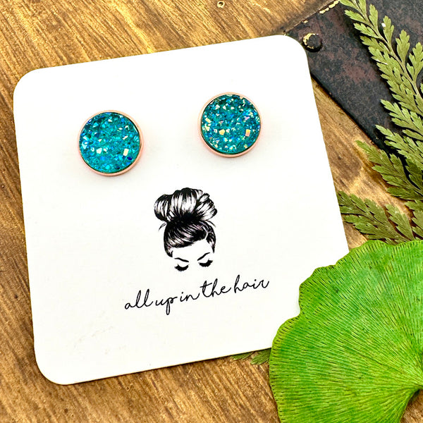 All Up In The Hair | Online Accessory Boutique Located in Mooresville, NC | Two Aquamarine Druzy set in a rose gold stud setting. The earrings are on a white, All Up In The Hair branded packaging card. The card is laying on a wood background, alongside green leaves.