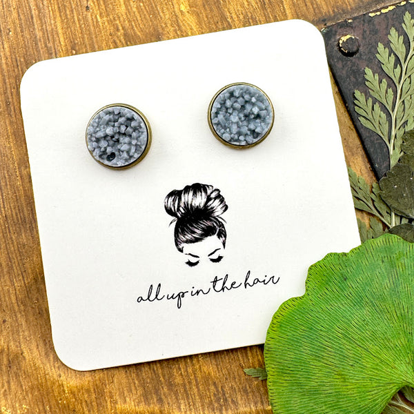 All Up In The Hair | Online Accessory Boutique Located in Mooresville, NC | Two ash grey druzies set in a bronze earring setting. The earrings are on a white, All Up In The Hair branded packaging card. The card is laying on a wood background next to green leaves.