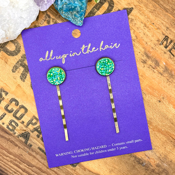 All Up In The Hair | Online Accessory Boutique Located in Mooresville, NC | An All Up In The Hair branded packaging card on a grey background, surrounded by colorful glitter. On the card is two aventurine druzy bobby pins.