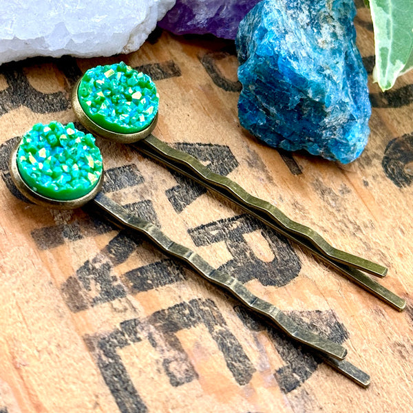 All Up In The Hair | Online Accessory Boutique Located in Mooresville, NC | Side view of our Aventurine Druzy Bobby Pins laying on a wood background with black lettering. At the top of the image are crystals and ivy leaves.