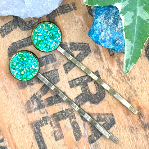 All Up In The Hair | Online Accessory Boutique Located in Mooresville, NC | Two green druzy bobby pins laying diagonally on a grey background, surrounded by colorful glitter.