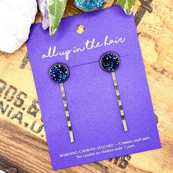 All Up In The Hair | Online Accessory Boutique Located in Mooresville, NC | An All Up In The Hair branded packaging card laying on a grey background, surrounded by colorful glitter. On the card is two azurite druzy bobby pins.