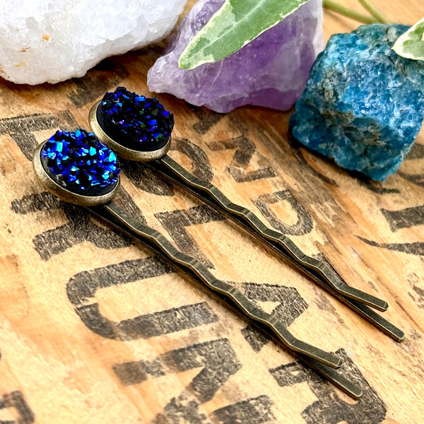 All Up In The Hair | Online Accessory Boutique Located in Mooresville, NC | Side view of our Azurite Druzy Bobby Pins on a wood background with black lettering. There are crystals and ivy leaves at the top of the image.