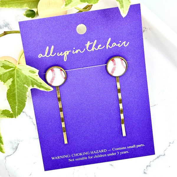 All Up In The Hair | Online Accessory Boutique Located in Mooresville, NC | An indigo colored, All Up In The Hair branded packaging card laying on a gray background, surrounded by colorful glitter. On the card is two baseball bobby pins.