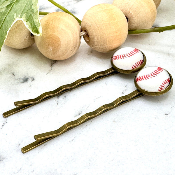 All Up In The Hair | Online Accessory Boutique Located in Mooresville, NC | Side view of two Baseball Bobby Pins on a white marble background. There is a wood bead garland and ivy leaves next to the bobby pins.