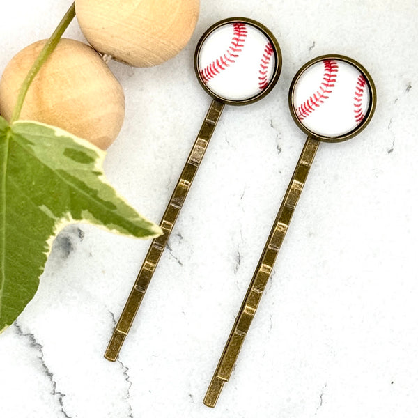 All Up In The Hair | Online Accessory Boutique Located in Mooresville, NC | Two Baseball Bobby Pins on a white marble background. Next to the bobby pins is a wood bead garland and ivy leaves.