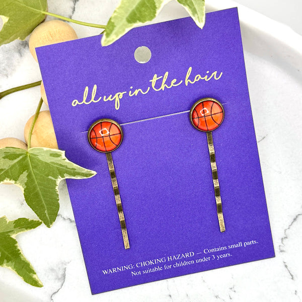 All Up In The Hair | Online Accessory Boutique Located in Mooresville, NC | Two Basketball Bobby Pins on an indigo colored, All Up In The Hair branded packaging card. The card is laying on a white marble background. There is a wood bead garland and ivy leaves next to the card.