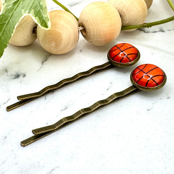 All Up In The Hair | Online Accessory Boutique Located in Mooresville, NC | Side view of two Basketball Bobby Pins on a white marble background. There is a wood bead garland and ivy leaves next to the bobby pins.