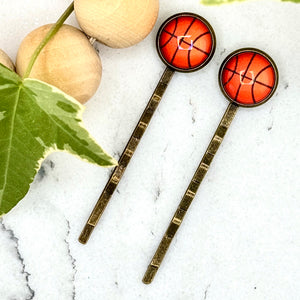 All Up In The Hair | Online Accessory Boutique Located in Mooresville, NC | Two Basketball Bobby Pins on a white marble background. Next to the bobby pins is a wood bead garland and ivy leaves.