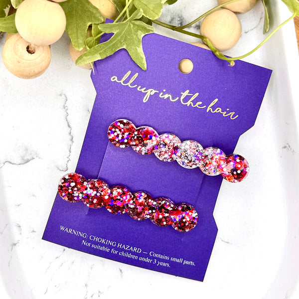 All Up In The Hair | Online Accessory Boutique Located in Mooresville, NC | Two Red Glitter Barrettes on an All Up In The Hair packaging. The card is laying on a gray background, surrounded by colorful glitter.