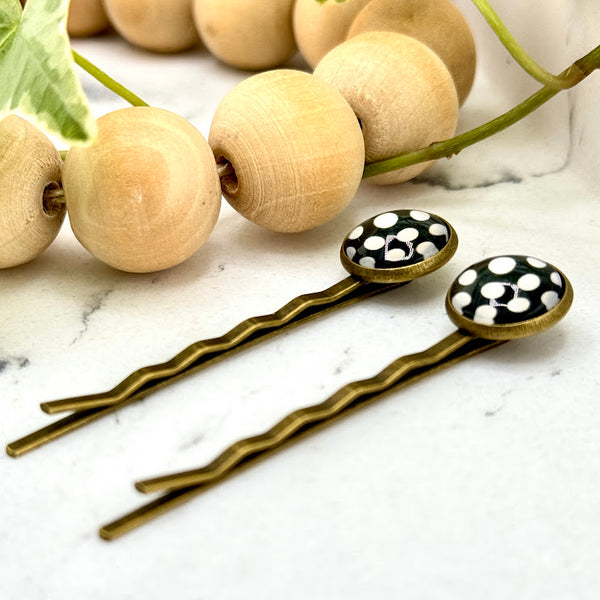 All Up In The Hair | Online Accessory Boutique Located in Mooresville, NC | Side view of two Big Polka Dot Bobby Pins on a white marble background. There is a wood bead garland and ivy leaves next to the bobby pins.
