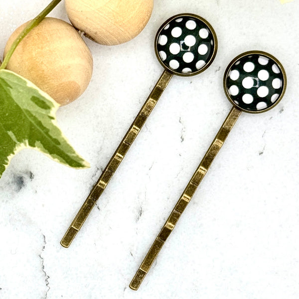 All Up In The Hair | Online Accessory Boutique Located in Mooresville, NC | Two bobby pins with a black cabochon with large white polka dots laying on a white marble background. There is a wood bead garland and ivy leaves next to the bobby pins.