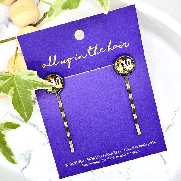 All Up In The Hair | Online Accessory Boutique Located in Mooresville, NC | Two Bird Of Paradise Bobby Pins on an indigo colored, All Up In The Hair branded packaging card. The card is laying on a white marble background. There is a wood bead garland and ivy leaves next to the card.
