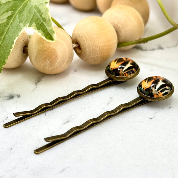 All Up In The Hair | Online Accessory Boutique Located in Mooresville, NC | Side view of our Bird Of Paradise Bobby Pins laying on a white marble background. There is a wood bead garland and ivy leaves next to the bobby pins.