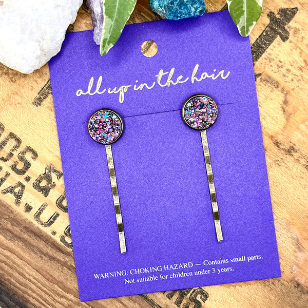 All Up In The Hair | Online Accessory Boutique Located in Mooresville, NC | An All Up In The Hair branded packaging card laying on a grey background, surrounded by colorful glitter. On the card is two bismuth druzy bobby pins.