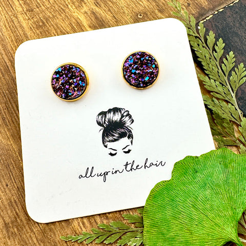 All Up In The Hair | Online Accessory Boutique Located in Mooresville, NC | Two rainbow colored Bismuth Druzy set in a gold stud setting. The earrings are on a white, All Up In The Hair branded card, laying on a wood background alongside green leaves.