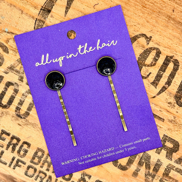 All Up In The Hair | Online Accessory Boutique Located in Mooresville, NC | Two Black Mermaid Bobby Pins on an All Up In The Hair branded, indigo colored, packaging card. The card is laying on a wood background with black lettering.