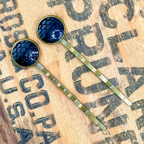 All Up In The Hair | Online Accessory Boutique Located in Mooresville, NC | Two Black Mermaid Bobby Pins laying diagonally on a wood background with black lettering.
